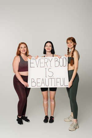 Sportswoman with vitiligo holding placard with every body is beautiful lettering near friends on...