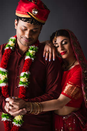Indian man in turban holding hands with happy bride in traditional headscarf isolated on gray