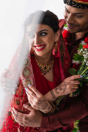 happy indian man in turban hugging smiling bride with mehndi holding flowers on whiteの写真素材