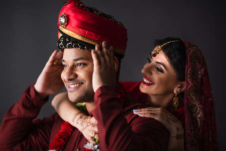 cheerful indian bride hugging bridegroom in turban isolated on grayの写真素材