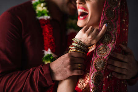 partial view of blurred indian man in turban holding hand of happy bride in traditional headscarf isolated on grayの写真素材