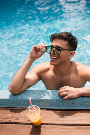 High angle view of man in sunglasses smiling while relaxing in swimming pool near orange juice
