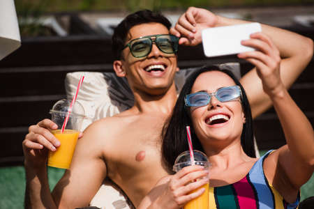 Happy woman taking selfie on smartphone near blurred shirtless boyfriend with orange juice on deck chairの写真素材
