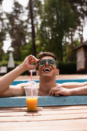 Positive man holding sunglasses near blurred orange juice on poolsideの写真素材
