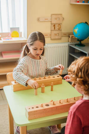girl playing game with wooden element near blurred kid in montessori school