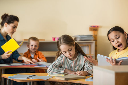 asian girl reading book near blurred teacher and children writing in notebooks during lessonの写真素材