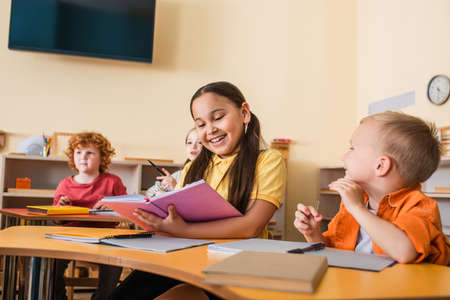 happy asian girl reading book near friends during lesson in montessori schoolの写真素材