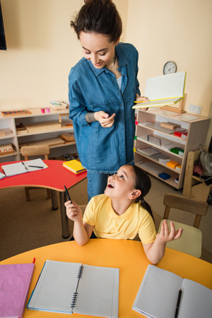 high angle view of amazed asian girl talking to smiling teacher in montessori schoolの写真素材