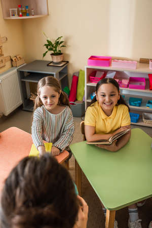 cheerful multiethnic girls looking at teacher on blurred foreground during lesson