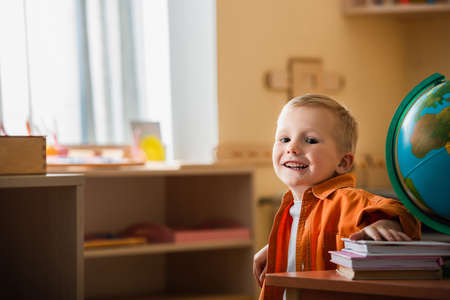happy boy smiling at camera near globe and books in montessori schoolの写真素材