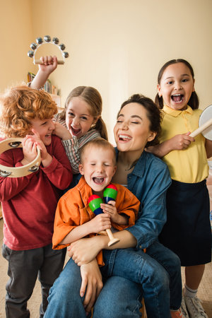 excited teacher laughing while multiethnic children playing musical instrumentsの写真素材