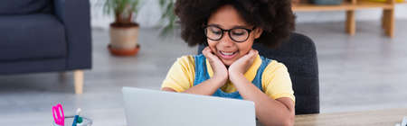 Happy african american kid looking at blurred laptop near stationery at home, banner