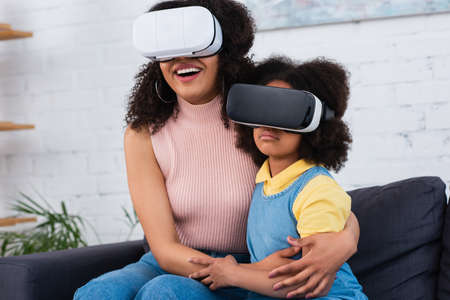 Smiling african american mother hugging daughter in vr headset at homeの写真素材