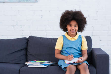 African american girl smiling at camera while doing schoolwork on couch