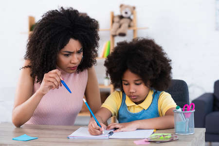 Angry african american mother pointing with pen near daughter writing on notebook at homeの写真素材