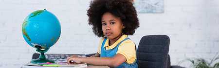 African american girl looking at camera near stationery and globe on table, bannerの写真素材