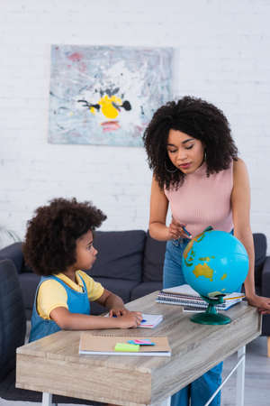 African american mother pointing at globe near daughter with notebooks