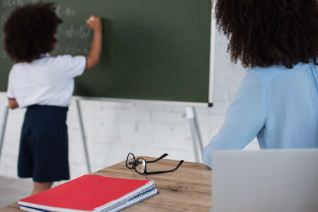 African american teacher sitting near laptop and blurred pupil writing on chalkboardの写真素材