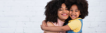Smiling african american kid embracing mother with closed eyes at home, bannerの写真素材