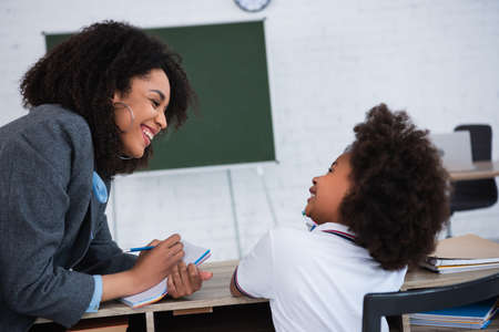 Smiling african american teacher writing on notebook near pupil in classroom
