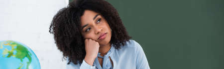 Pensive african american teacher looking away near blurred globe in classroom, banner
