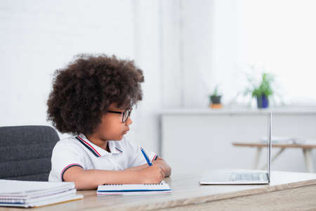 Side view of african american schoolgirl with pen and notebook near laptop in classroom