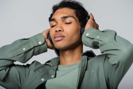 young african american man touching headphones while listening music with closed eyes isolated on grayの写真素材