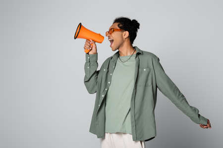 trendy african american guy shouting in orange megaphone isolated on gray