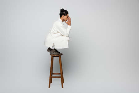 side view of african american guy in white clothes posing with praying hands on stool on gray backgroundの写真素材