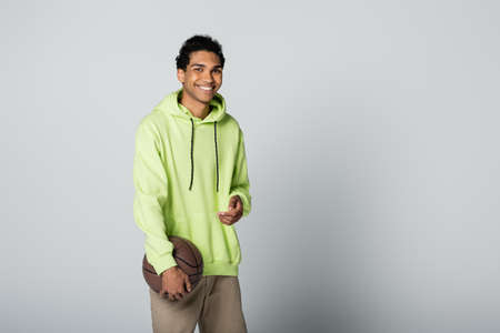 stylish african american man in green hoodie smiling at camera while standing with basketball on...