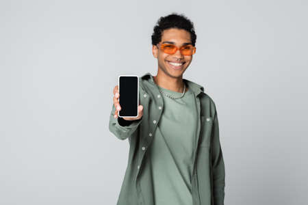 blurred african american man smiling while showing smartphone with blank screen isolated on grayの写真素材
