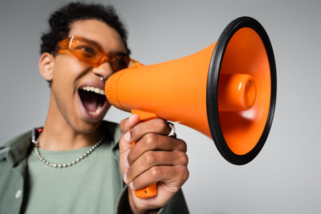 blurred african american guy in orange eyeglasses screaming in loudspeaker isolated on grayの写真素材