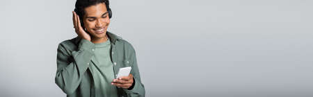 joyful african american man looking at cellphone while listening music isolated on gray, banner