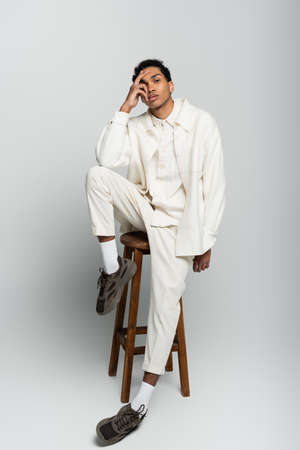 african american man in trendy clothes sitting on high stool with hand on face on gray background