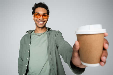 cheerful and stylish african american guy holding blurred paper cup isolated on grayの写真素材