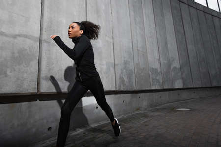 Young african american sportswoman running near concrete wall with shadow on urban streetの写真素材