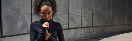 African american sportswoman looking at camera while zipping sports jacket outdoors, banner