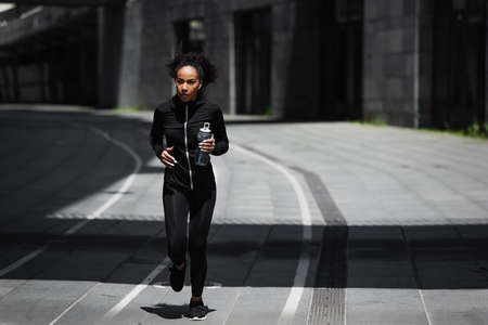 African american runner with sports bottle listening music in earphones on urban streetの写真素材