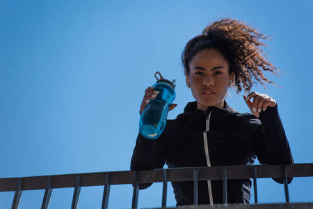 Low angle view of african american sportswoman in earphones holding sports bottle on bridge and...