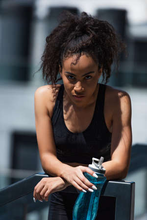 African american sportswoman holding sports bottle and looking at camera near railing