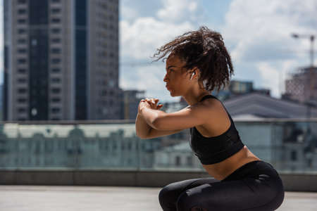 Side view of african american sportswoman in earphone doing squat on urban streetの写真素材