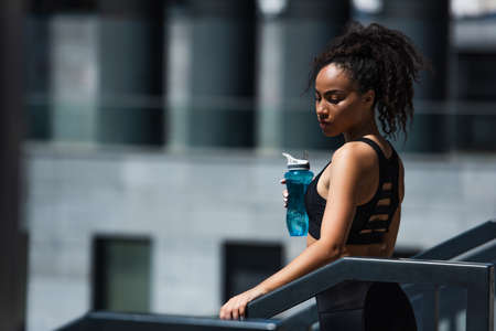 African american sportswoman in black top holding sports bottle near railing outdoorsの写真素材