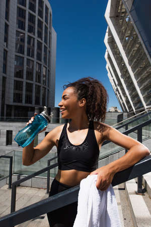 Side view of happy african american sportswoman with towel smiling while holding water near railing