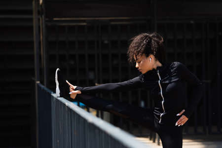 Side view of african american sportswoman in wireless earphone stretching near bridge on urban...