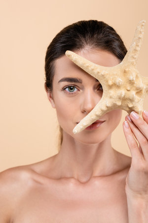 charming woman with naked shoulders obscuring face with starfish isolated on beige, beauty conceptの写真素材