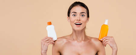amazed woman showing bottles of sunblock while looking at camera isolated on beige, banner