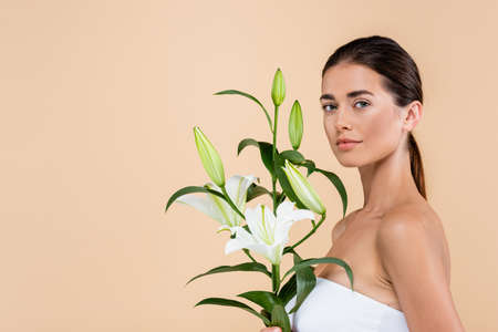 pretty woman looking at camera near white lily flowers isolated on beige, beauty conceptの写真素材