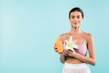 young woman with bottle of sunscreen and white lily looking at camera isolated on blue