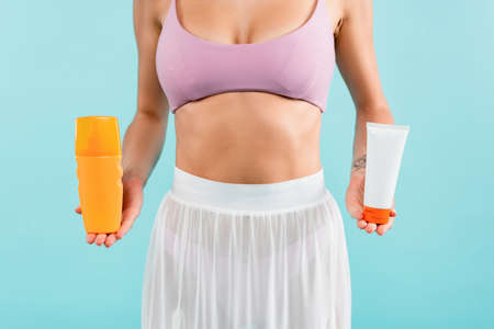 cropped view of woman in swimsuit and pareo holding tube and bottle of sunscreen isolated on blue