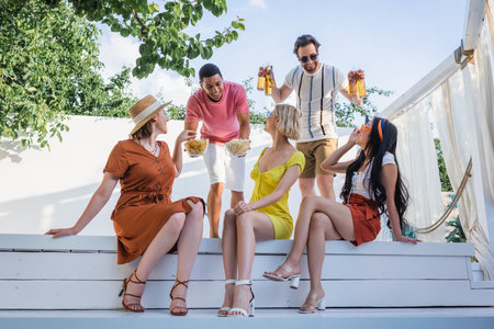 Positive multiethnic men holding beer and food near friends in patio